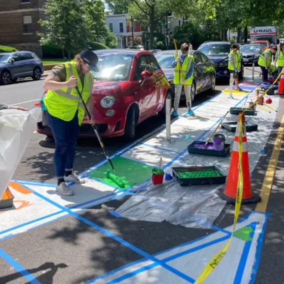 Volunteers in reflective vests paint colorful designs on a city street next to parked cars, using rollers, tape, and traffic cones to create a safer and more vibrant public space.