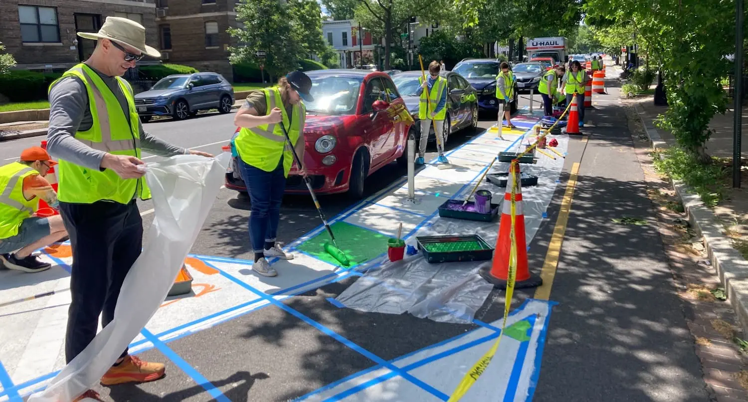 Volunteers in reflective vests paint colorful designs on a city street next to parked cars, using rollers, tape, and traffic cones to create a safer and more vibrant public space.