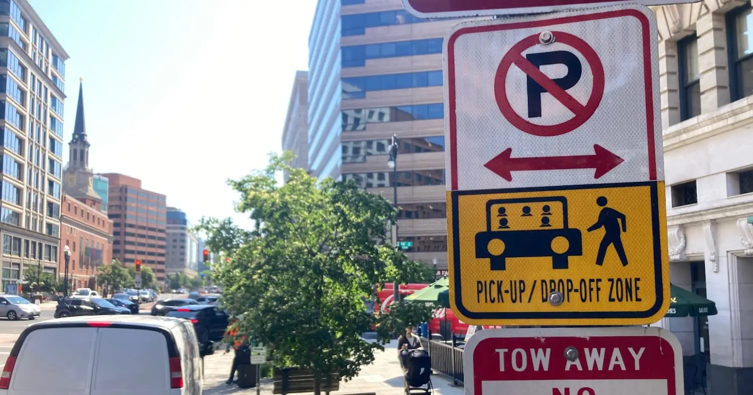 A street sign in a busy downtown area designating a no-parking pick-up and drop-off zone, with cars, pedestrians, and tall office buildings in the background.