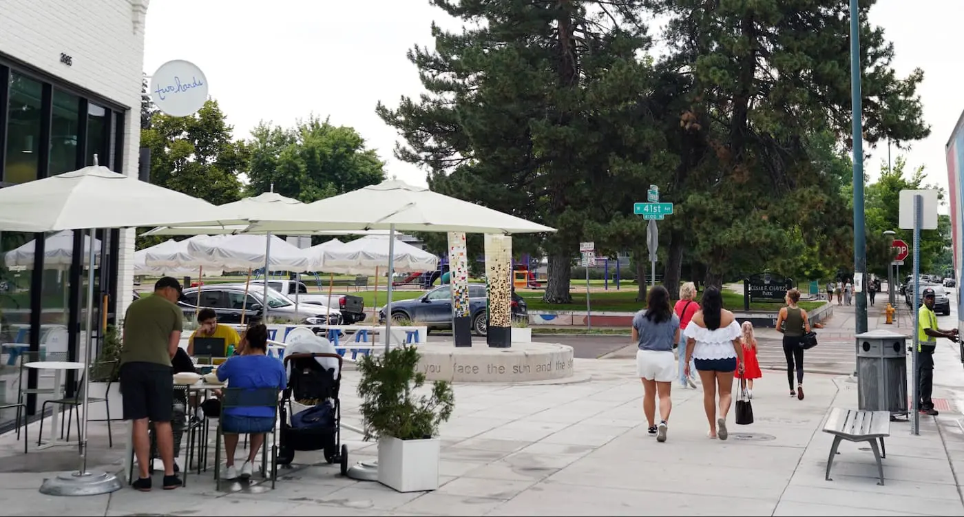A lively city street with outdoor café seating under umbrellas, families and friends walking along the sidewalk, and a nearby park with trees and playground equipment in the background. The scene shows a vibrant, walkable neighborhood space.
