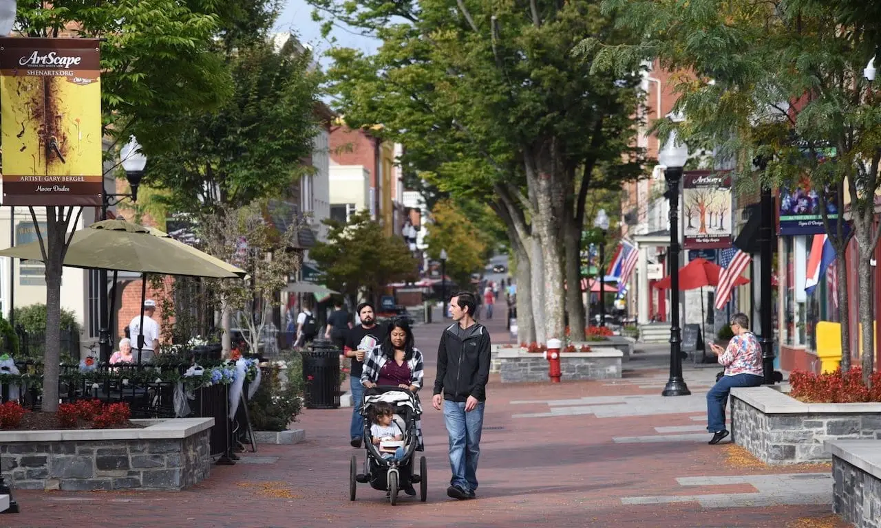 A tree-lined brick pedestrian street in Winchester, Virginia, with families walking, a woman pushing a stroller, outdoor café seating under umbrellas, storefronts with American flags, and people enjoying the lively, walkable downtown.