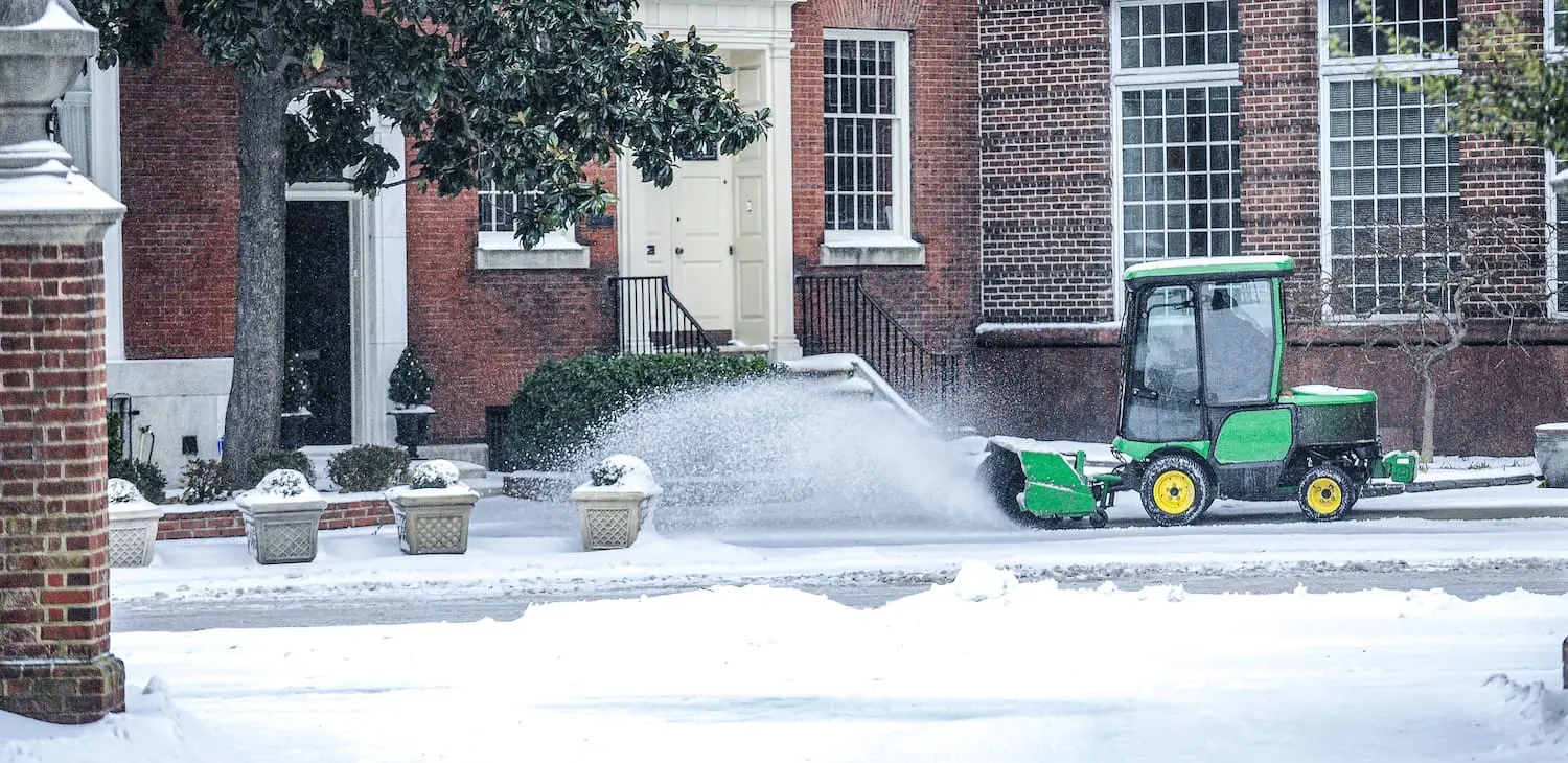 A small green snow plow clears snow from a city street in front of historic brick row buildings in Philadelphia.