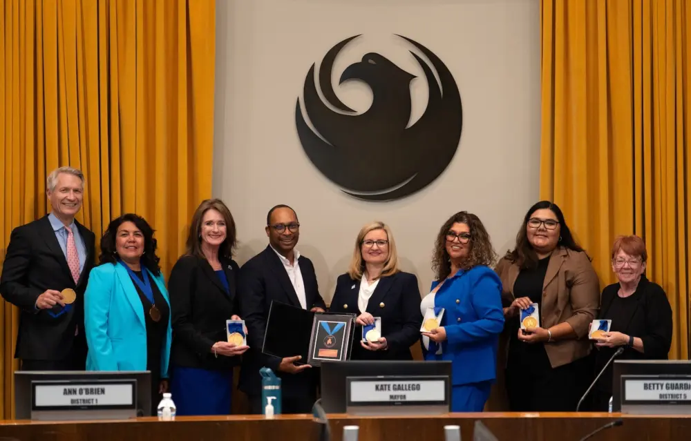 A group of eight people, including Mayor Kate Gallego, stand for a photograph behind a wooden council dais, during the presentation of an award. Seven of the attendees are holding circular gold medals, while a man in a black jacket (fourth from left) presents a framed plaque with a larger award to Mayor Gallego (center, wearing glasses). A large, stylized dark phoenix emblem is mounted on the wall behind them, flanked by gold curtains. Nameplates below identify council members Ann O’Brien and Betty Guardado, as well as Mayor Kate Gallego.