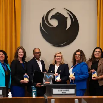 A group of eight people, including Mayor Kate Gallego, stand for a photograph behind a wooden council dais, during the presentation of an award. Seven of the attendees are holding circular gold medals, while a man in a black jacket (fourth from left) presents a framed plaque with a larger award to Mayor Gallego (center, wearing glasses). A large, stylized dark phoenix emblem is mounted on the wall behind them, flanked by gold curtains. Nameplates below identify council members Ann O’Brien and Betty Guardado, as well as Mayor Kate Gallego.