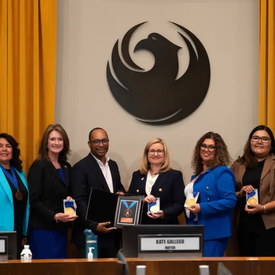 A group of eight people, including Mayor Kate Gallego, stand for a photograph behind a wooden council dais, during the presentation of an award. Seven of the attendees are holding circular gold medals, while a man in a black jacket (fourth from left) presents a framed plaque with a larger award to Mayor Gallego (center, wearing glasses). A large, stylized dark phoenix emblem is mounted on the wall behind them, flanked by gold curtains. Nameplates below identify council members Ann O’Brien and Betty Guardado, as well as Mayor Kate Gallego.