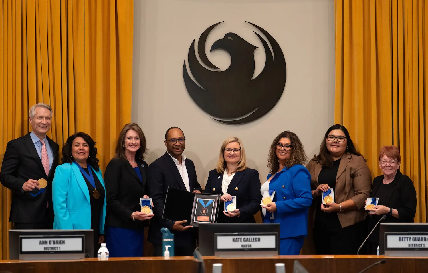 A group of eight people, including Mayor Kate Gallego, stand for a photograph behind a wooden council dais, during the presentation of an award. Seven of the attendees are holding circular gold medals, while a man in a black jacket (fourth from left) presents a framed plaque with a larger award to Mayor Gallego (center, wearing glasses). A large, stylized dark phoenix emblem is mounted on the wall behind them, flanked by gold curtains. Nameplates below identify council members Ann O’Brien and Betty Guardado, as well as Mayor Kate Gallego.