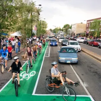 Cyclists ride down a green-painted, clearly marked bike lane on a busy downtown street, including one person on a recumbent bike in the foreground.Cyclists ride down a green-painted, clearly marked bike lane on a busy downtown street, including one person on a recumbent bike in the foreground. A line of cars travels in the adjacent vehicle lanes to the right, while pedestrians fill the sidewalk on the left near storefronts, street trees, and a festival-style tent. The scene shows a lively, mixed-use street where biking, driving, and walking happen side by side.
