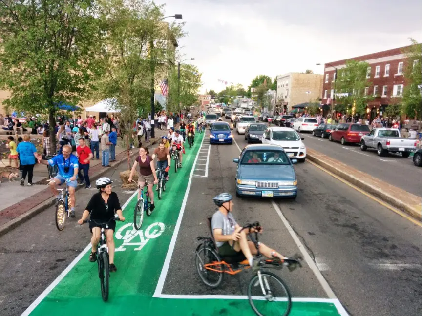 Cyclists ride down a green-painted, clearly marked bike lane on a busy downtown street, including one person on a recumbent bike in the foreground.Cyclists ride down a green-painted, clearly marked bike lane on a busy downtown street, including one person on a recumbent bike in the foreground. A line of cars travels in the adjacent vehicle lanes to the right, while pedestrians fill the sidewalk on the left near storefronts, street trees, and a festival-style tent. The scene shows a lively, mixed-use street where biking, driving, and walking happen side by side.