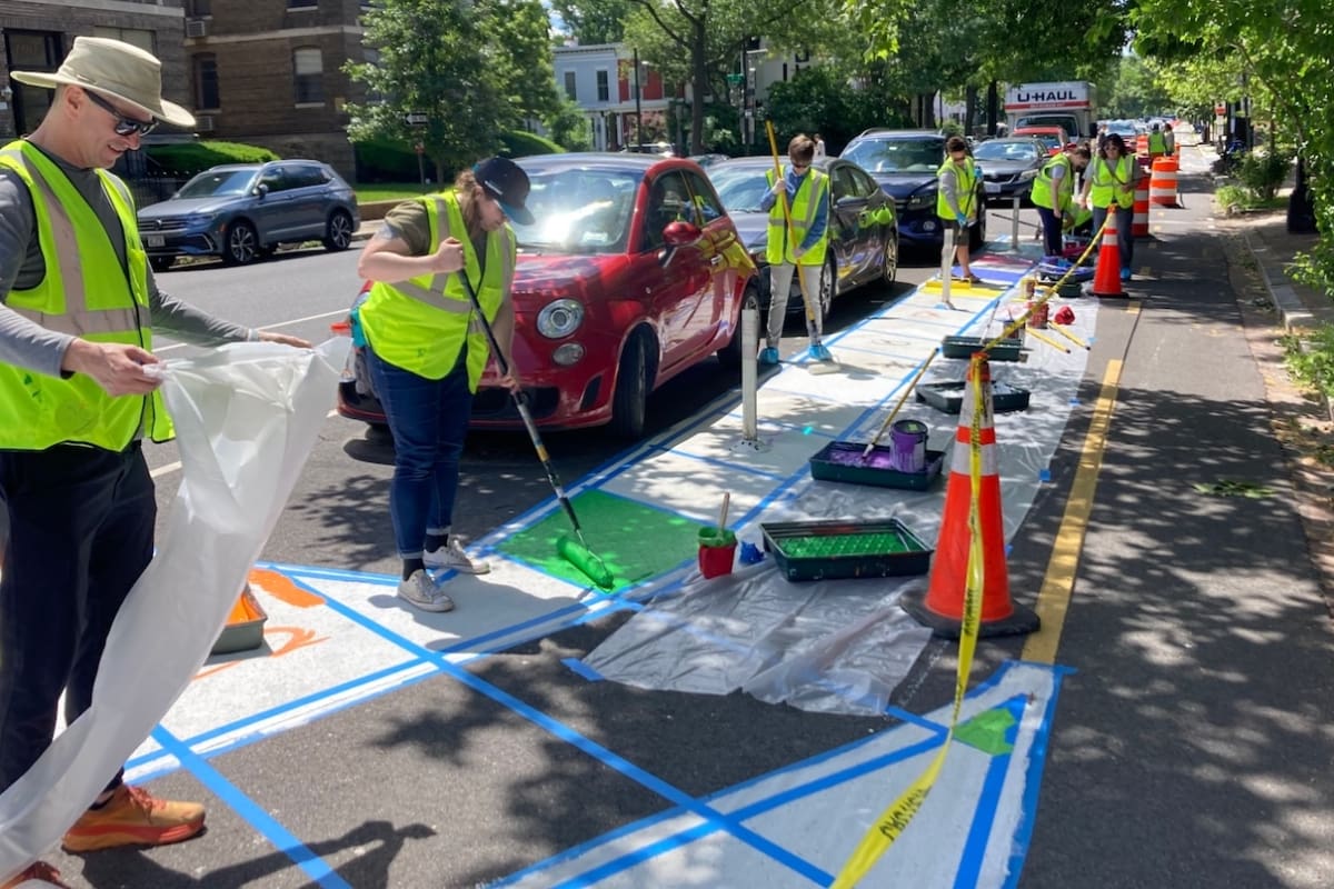 Volunteers in reflective vests paint colorful designs on a city street next to parked cars, using rollers, tape, and traffic cones to create a safer and more vibrant public space.